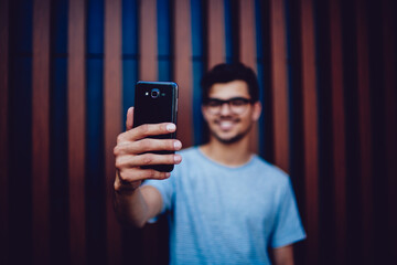 Selective focus on modern smartphone in hands of hipster guy making selfie for update profile picture in networks, blurred male teenager in t-shirt smiling taking photo using mobile phone camera