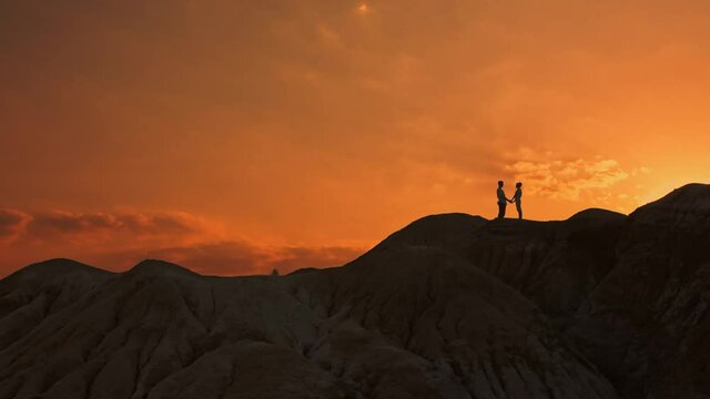 Against the backdrop of a bright orange sky at the top of the mountain with stony slopes in the general plan are two silhouettes of a couple in love, a man and a woman, they hold hands and look at