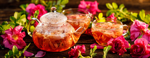 Rose tea infusion of rose petals in a glass teapot and cups among fresh rose flowers on a wooden table