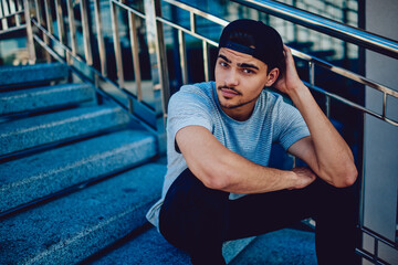 Portrait of handsome young man dressed in casual street outfit sitting on stairs looking at camera,...