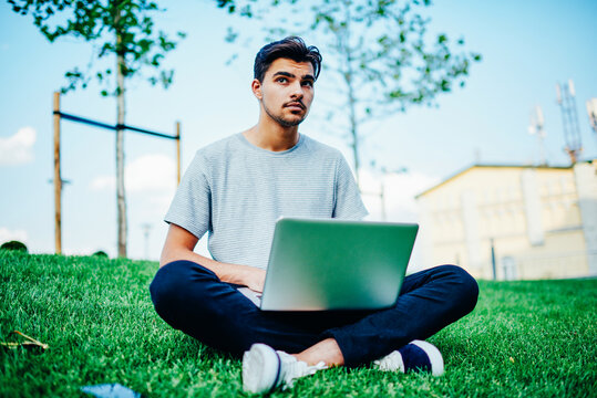 Pondering smart young student dressed in casual outfit thinking on writing coursework on modern laptop computer sitting on green grass and using free 4G internet on campus territory