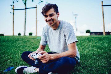 Portrait of cheerful young guy sitting on green lawn enjoying weekends holding vintage camera for taking pictures, handsome male hipster having fun during leisure spending time on photography