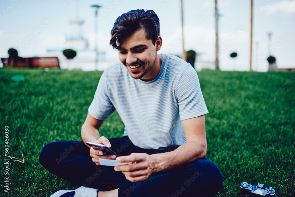 Poster cheerful hipster guy browsing website on smartphone excited with using promo code from card getting 