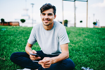 Half length portrait of happy hipster student smiling at camera while dialing phone number on smartphone.Positive male blogger chatting online on cellular via internet sitting on geen grass in campus