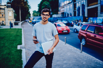 Portrait of casually dressed male student standing on street with smartphone texting SMS while waiting for friend, hipster guy share photos in social networks walking in town holding laptop