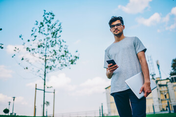 Full length portrait of student in eyewear strolling on street while updating profile on smartphone via free 4g internet.Young blogger with phone and laptop in hand looking at camera walking outdoors