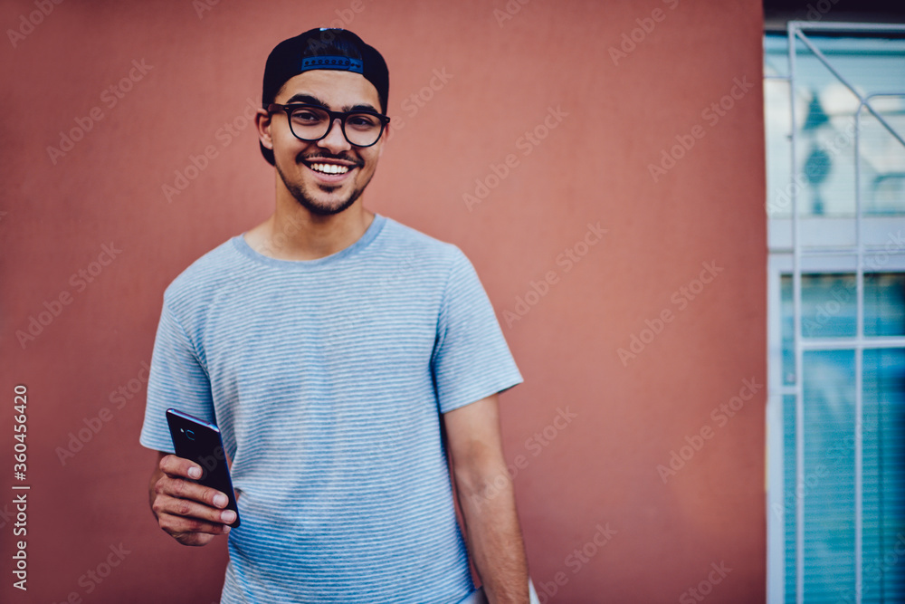 Poster portrait of cheerful hipster guy in trendy eyeglasses using mobile for sending messages standing on 