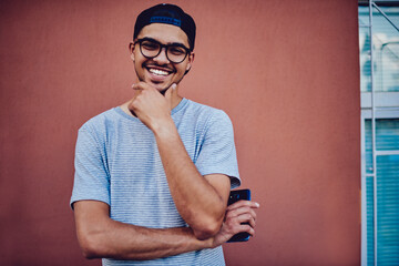 Portrait of cheerful hipster guy in trendy street look excited with idea of free time waiting for call on promotional background, smiling blogger dressed in textile t-shirt and cap posing on wall