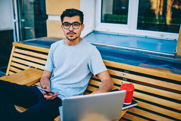 Portrait of handsome young man in eyewear looking at camera sitting on wooden bench with modern...