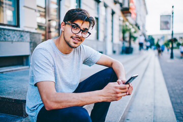 Portrait of successful student in eye glasses smiling at camera while updating profile in social networks on smartphone device using free 4G internet connection sitting on street in urban setting