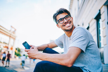 Happy young man in eye glasses laughing while holding smartphone in hand sitting outdoors in urban setting.Positive hipster blogger with mobile phone having fun on street in downtown