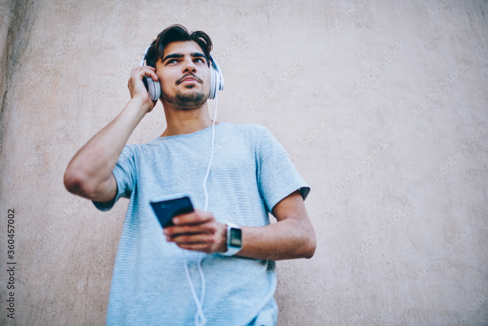 Poster cheerful hipster guy in modern headphone listening new song from internet radio website using smartp