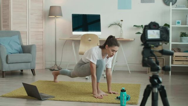 Wide shot of young female yoga coach wearing grey leggings and white t-shirt is standing on mat in front of camera and doing set of body stretching exercises