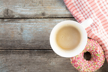 A pink doughnut on a wooden planks background with a cup of coffee with milk and a napkin on a vintage wood background with copy space.