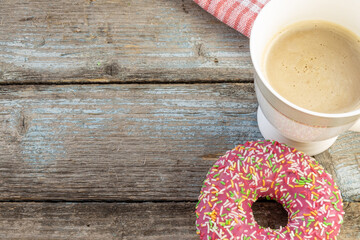 A pink doughnut on a wooden planks background with a cup of coffee with milk and a napkin on a vintage wood background with copy space.