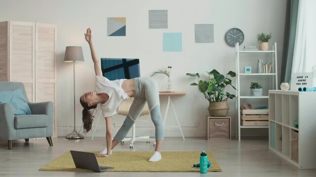 Wide shot of young female yoga tutor wearing grey leggings and white t-shirt is standing on mat at laptop and stretching her body