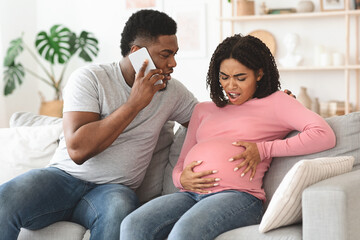 Pregnant black woman suffering, husband calling doctor, home interior