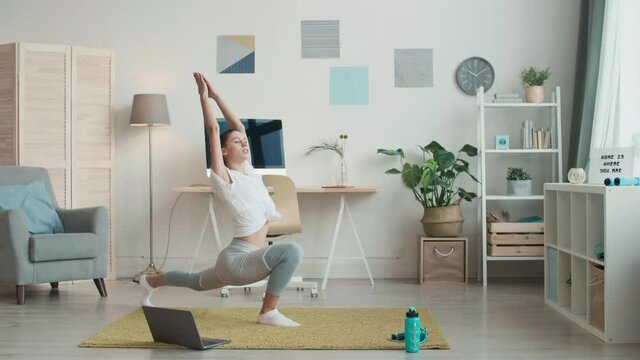 Time lapse of slim female yoga tutor wearing grey leggings and white t-shirt is standing on mat in front of laptop and doing set of stretching exercises