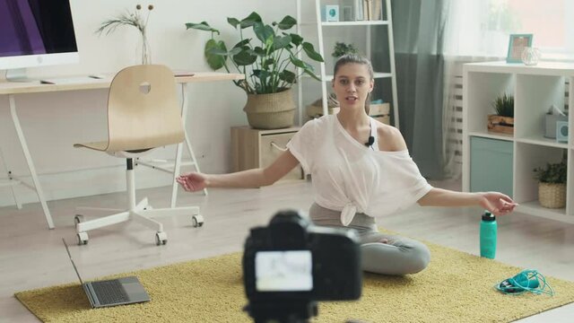 Panning of young female yoga tutor taking online yoga classes. Sportswoman is sitting on mat, looking at camera and telling about training purpose