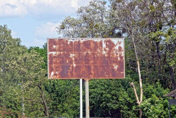 one old iron billboard in brown rust on the street against a background of green trees and sky