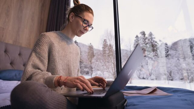 Portrait of attractive freelancer girl in glasses with a laptop on the bed in an eco-hotel with transparent walls behind which there is a winter forest. Travel and work concept with free schedule.