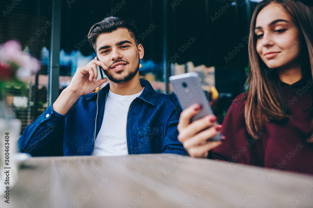Poster portrait of handsome male talking on mobile phone and looking at camera while her girlfriend using s