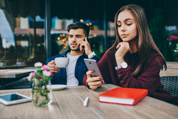 Pensive teen hipster girl reading notification on mobile phone sitting with handsome boyfriend at cafe terrace while he having telephone conversation satisfied with tariffs for internet connection