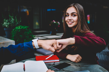 Portrait of charming young female student greeting with male colleague bumping fist sitting on rettace, cute hipster girl looking at camera while knuckles with guy satisfied with agreement of project