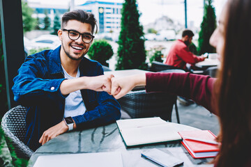 Cheerful hipster guy in eyewear bumping fists in sign of greeting with female friend having meeting on cafe terrace, cheerful young man enjoying completing successful project with partner knuckles