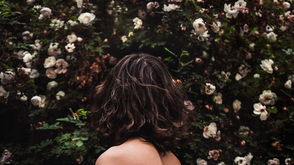 A girl with bare shoulders inhales the fragrance of garden flowers and stands with her back to the camera. Meditation against a living green wall with flowers