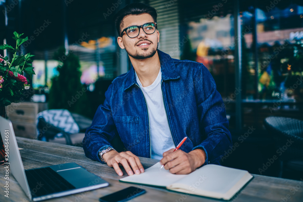 Poster pensive bearded guy in spectacles writing in notepad ideas for publication in blog sitting with lapt