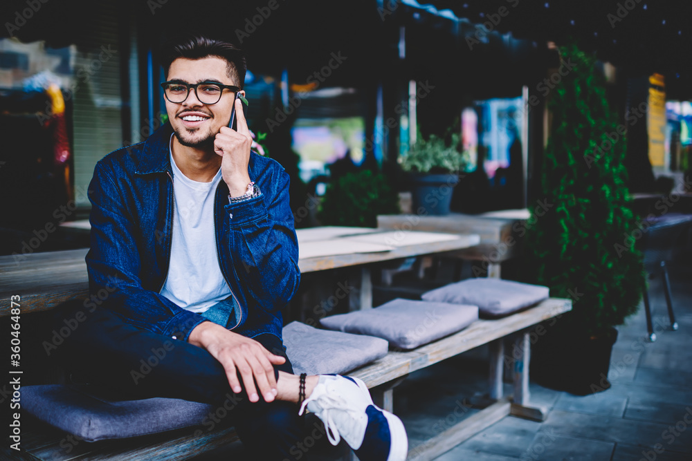 Poster portrait of cheerful hipster guy in spectacles talking on mobile with friend satisfied with tariffs 
