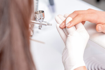 Manicure master is painting finger nails in beige color by airbrush in beauty salon
