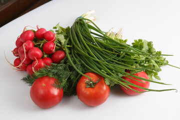 close up top view shot of a vegetable composition consisting of a bunch of radish, dill, parsley, green onions and ripe red tomatoes on a white background