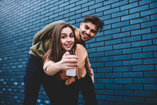Young hipster girl spending time with her boyfriend taking common pictures on smartphone camera, happy couple in love posing for selfie making funny images near copy space area for advertising