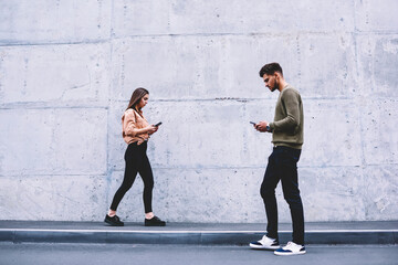 Young people walking on street near publicity area concentrated on technology spending time online, male and female hipsters strolling with smartphones addicted to social networks and digital devices