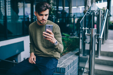 Pensive caucasian male using mobile phone for reading latest news from networks using 4G internet, concentrated blogger checking new feeds and sending mail via smartphone spending time outdoors