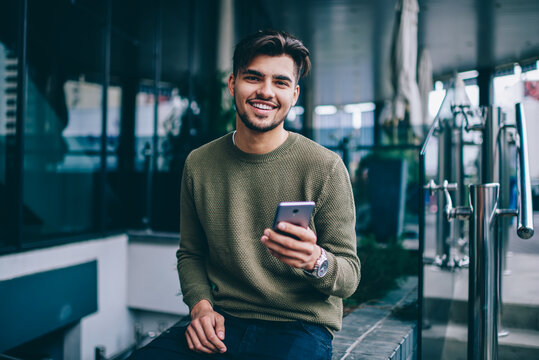 Portrait of cheerful handsome male using mobile phone and wireless internet for chatting and networking , young hipster guy looking at camera satisfied with good 4G connection for sending messages