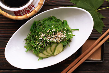 Chuka salad with cucumbers, sesame seeds and sauce, in a white plate, with a Japanese teapot, chopsticks and maple leaves, on a wooden background