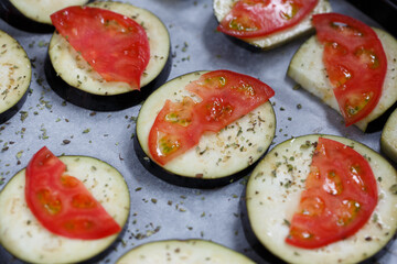 isolated filled frame close up shot of uncooked raw sliced aubergine eggplant with thin tomato slices on top sprinkled with dry green basil and oregano herbs, lying on a white baking paper background