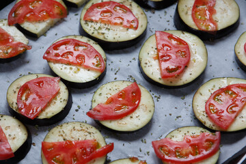 isolated filled frame close up shot of uncooked raw sliced aubergine eggplant with thin tomato slices on top sprinkled with dry green basil and oregano herbs, lying on a white baking paper background