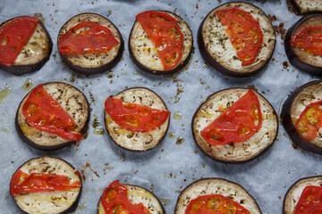 isolated filled frame close up shot of baked sliced aubergine eggplant with thin tomato slices on top, sprinkled with dry green basil and oregano herbs, lying on a white baking paper background