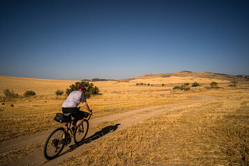 A man riding a gravel bike on Spanish farm tracks through fields of yellow grass with deep blue sky.