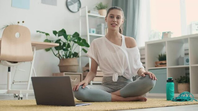 Lockdown of attractive female yoga coach is sitting on mat in front of laptop, looking at camera and beginning yoga practice by internet