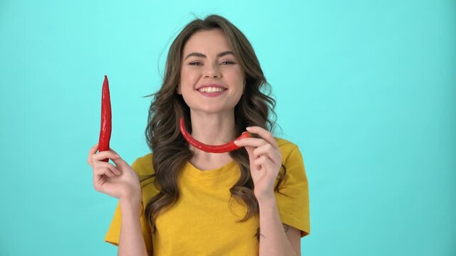 A positive nice young woman wearing a yellow t-shirt is posing with red chili peppers standing isolated over blue background