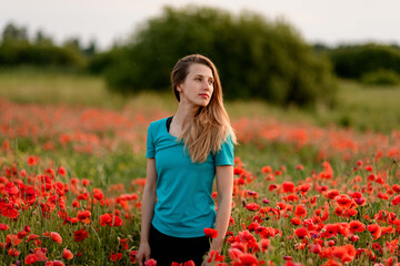 handsome young blond woman with long hair standing on field with red poppies and looks away.