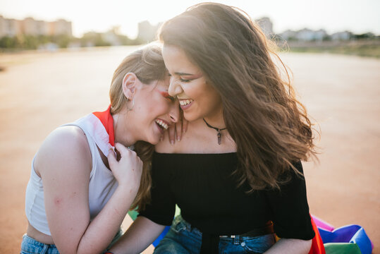 Two young girls sitting close and laughing at sunset with a rainbow flag of LGBT pride