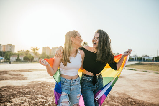 Two young girls (15-20 years old) walk around laughing with the LGBT Pride flag at sunset
