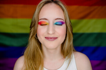 Portrait of young blonde girl smiling with colorful makeup against the background of the rainbow flag of gay pride