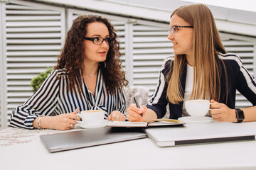 Two young beautiful smiling girl office workers discuss an important successful business project in a free environment in a cafe